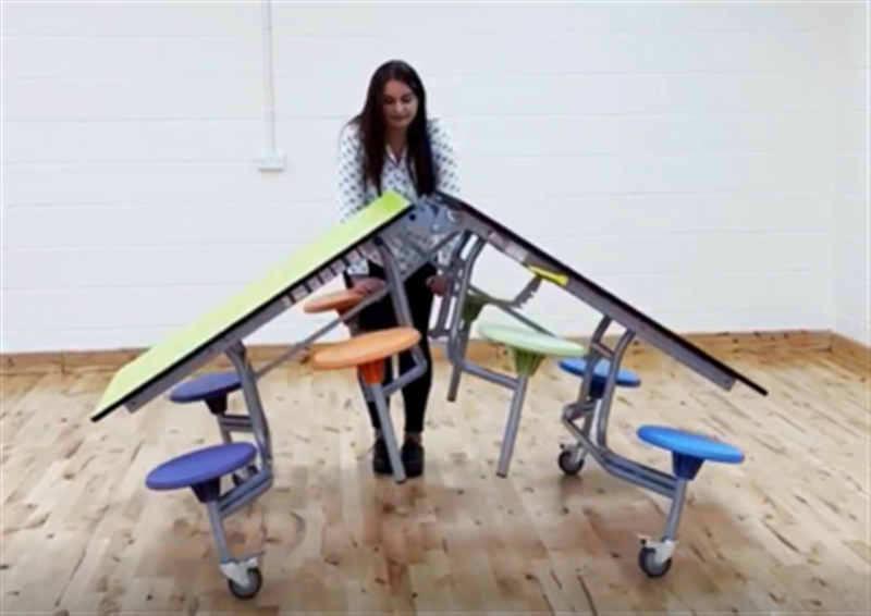A woman is unfolding an 8 seater school dining table. The table has a green top with purple, orange, blue and green seats. This is happening in a room with wooden flooring and white walls.